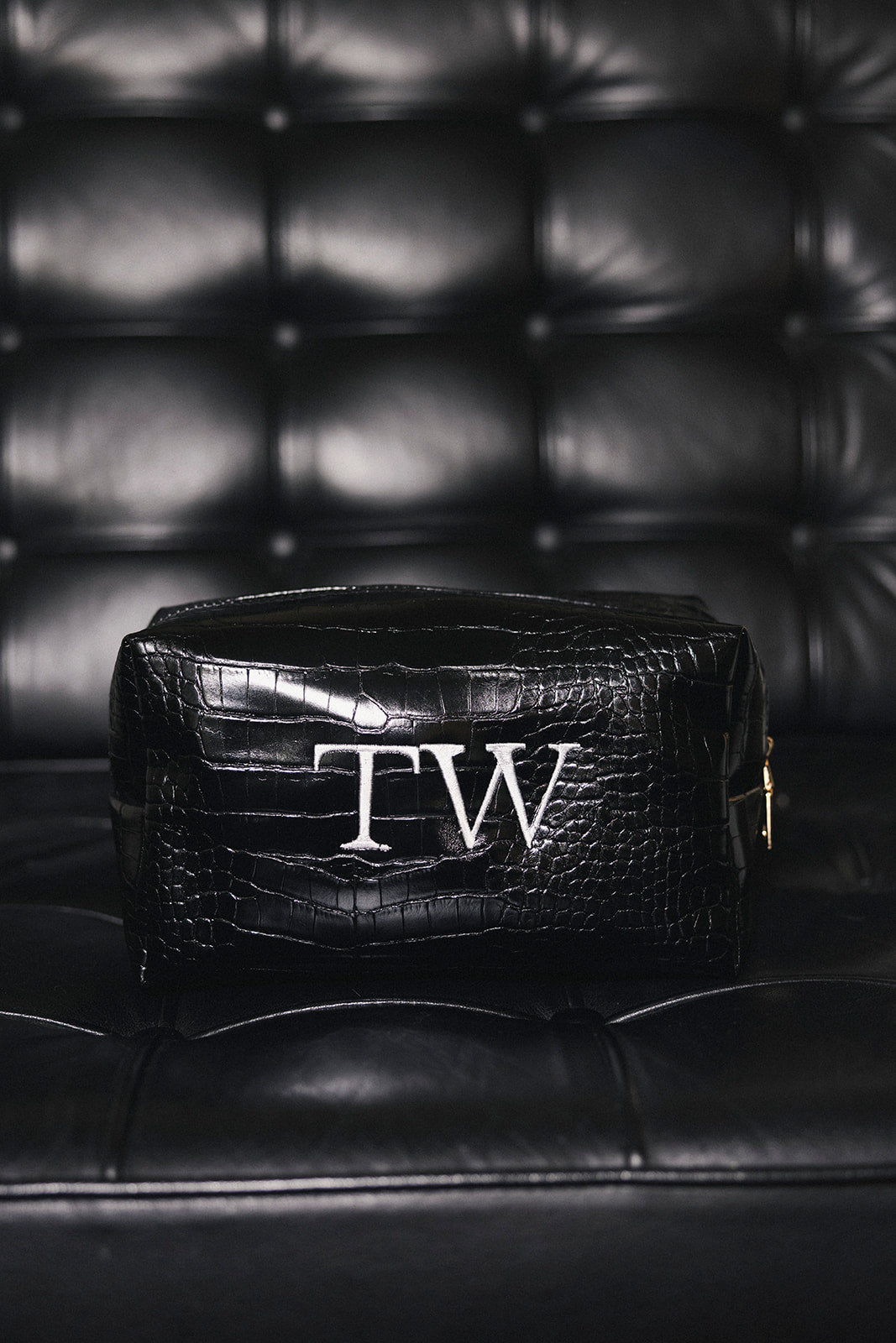 A black crocodile-embroidered leather cosmetic pouch with a white monogram 'TW' on the front, resting on a black leather tufted sofa.