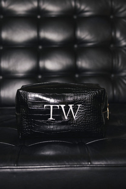 A black crocodile-embroidered leather cosmetic pouch with a white monogram 'TW' on the front, resting on a black leather tufted sofa.